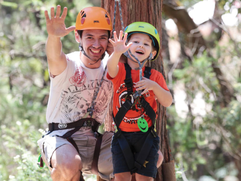 Parent and child in safety harness and helmet smiling and waving on a platform on the Nippers Tree Surfing course