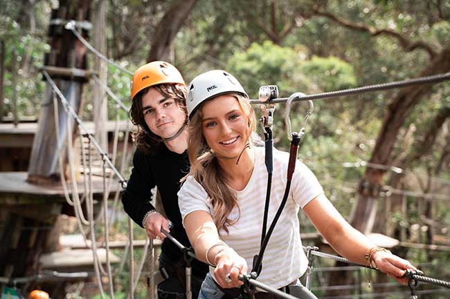 Two teens looking at camera smiling whilst crossing rope obstacle on Grand Tree Surfing course at Enchanted Adventure.