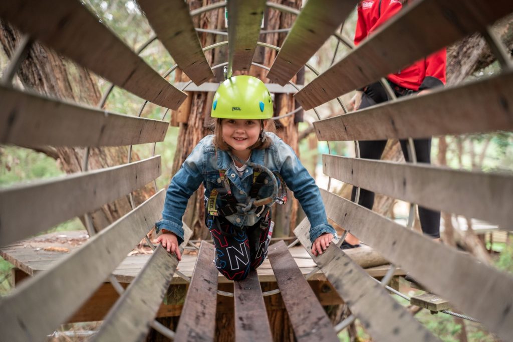 Young girl in helmet and harness crawling through a tunnel bridge on the Nippers Tree Surfing at Enchanted Adventure